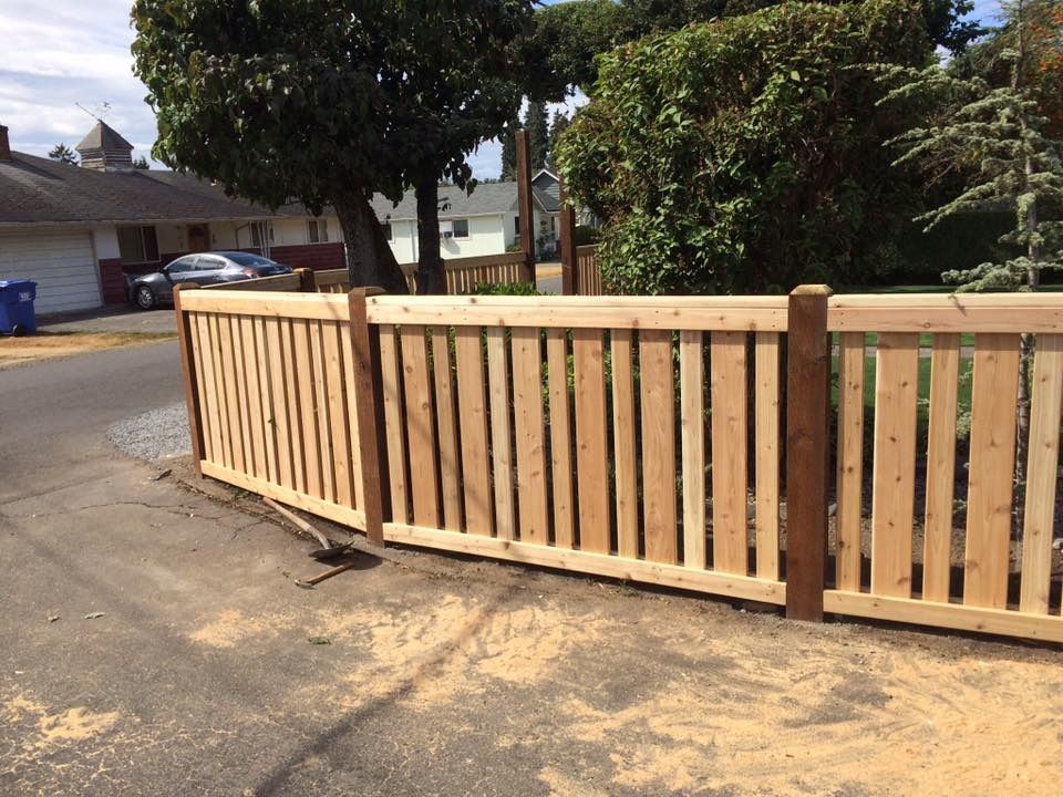 Wooden fence bordering a paved alley, with trees and houses in the background.