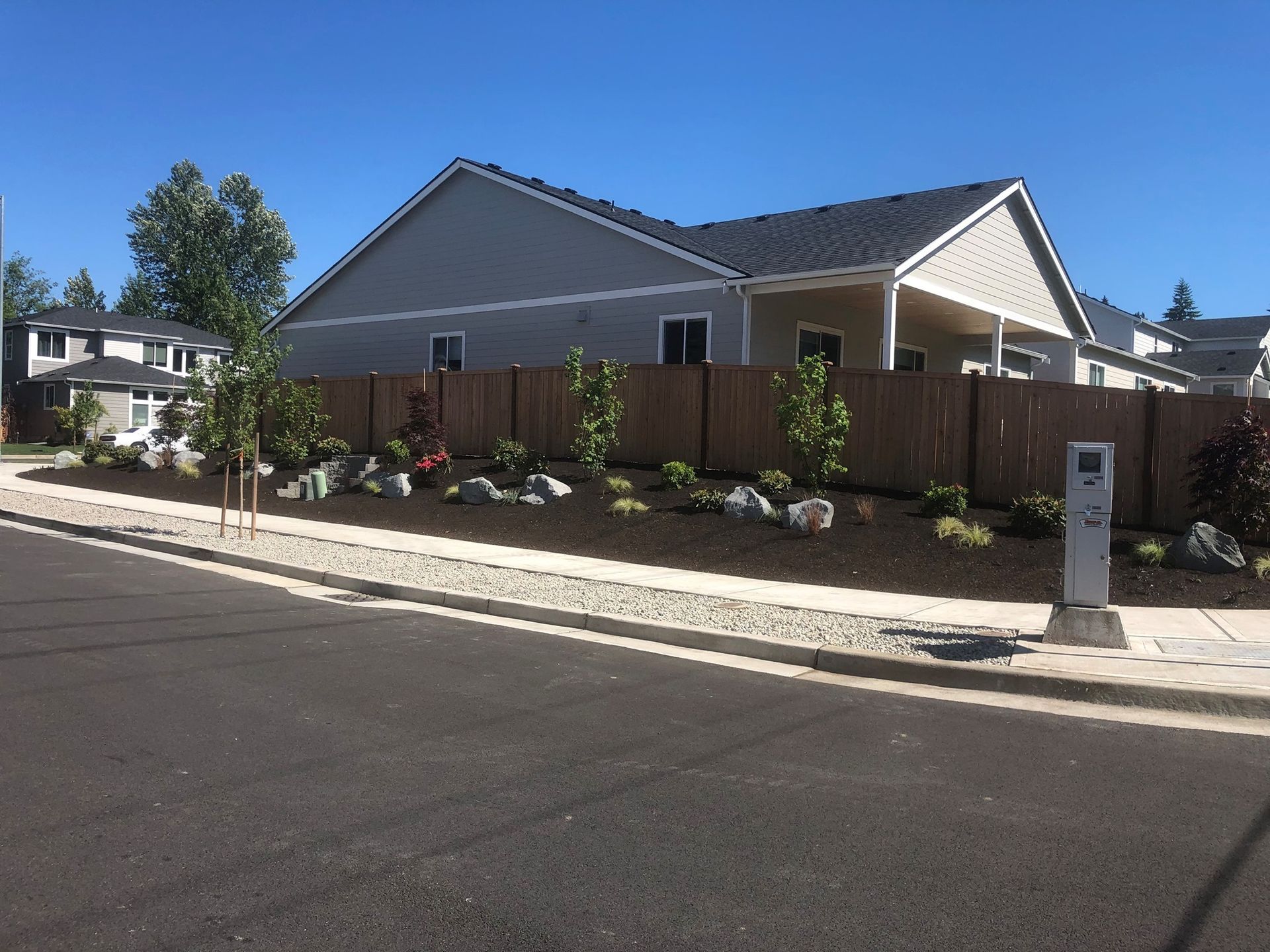 A house with a brown fence, landscaped with rocks and young trees along the edge of a road, on a sunny day.