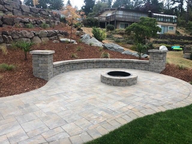 Stone patio with a fire pit, curved retaining wall, and two stone pillars; a hillside background.