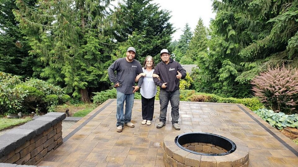 Three people stand on a brick patio with thumbs up. Lush green trees surround them. Fire pit in foreground.
