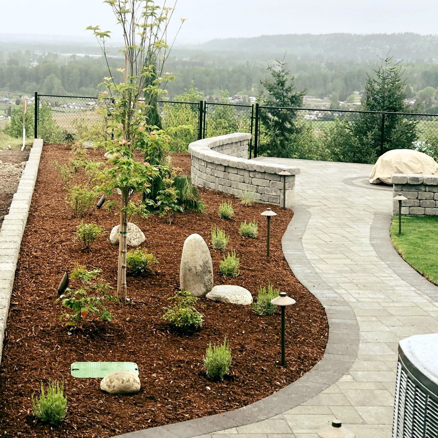 Stone patio with a garden bed, stone wall, and view of a valley. Brown mulch, trees, and pathway lights.