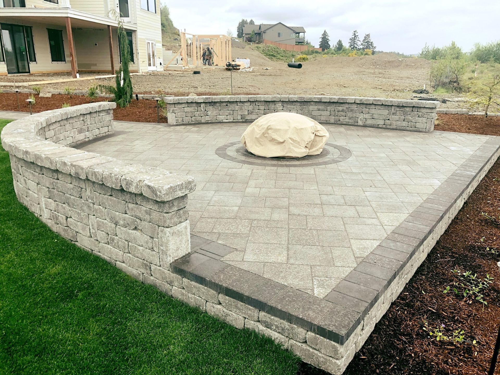 Stone patio with fire pit, curved seating wall, and rock in center. Green grass and construction in background.