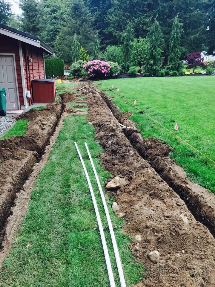 Trenches dug in a grassy yard, white pipes laid within, near a red building and garden.