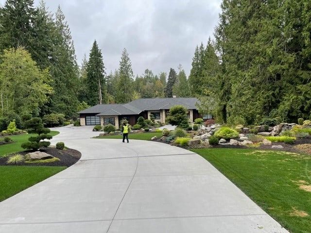 House with long driveway, landscaping, and a person standing on the driveway. Trees frame the house on a cloudy day.