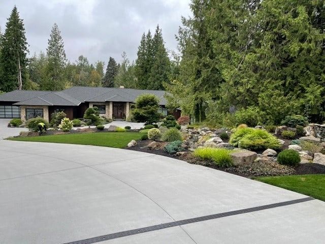 Low-slung house with curved driveway, landscaped with rocks, greenery, and trees. Overcast sky.