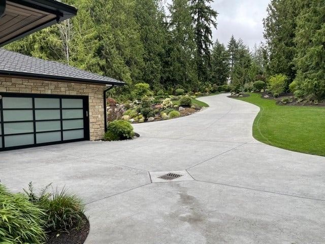 Concrete driveway curving up to a stone-clad garage with a glass door, surrounded by landscaping and trees.