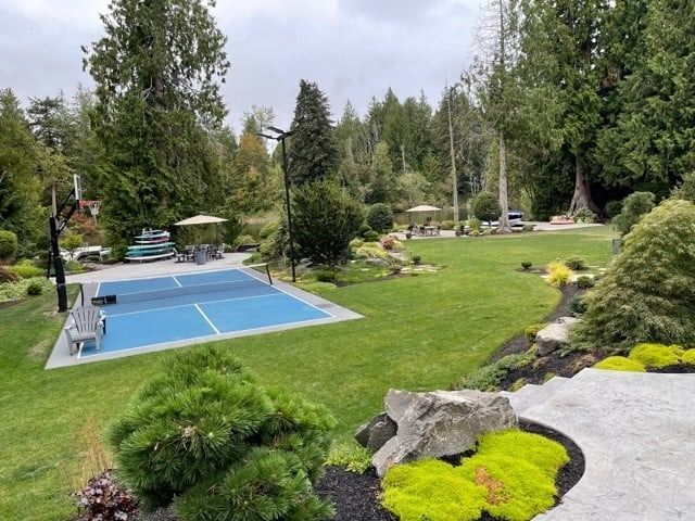 A lush green backyard with a blue pickleball court. Trees surround a grassy area.