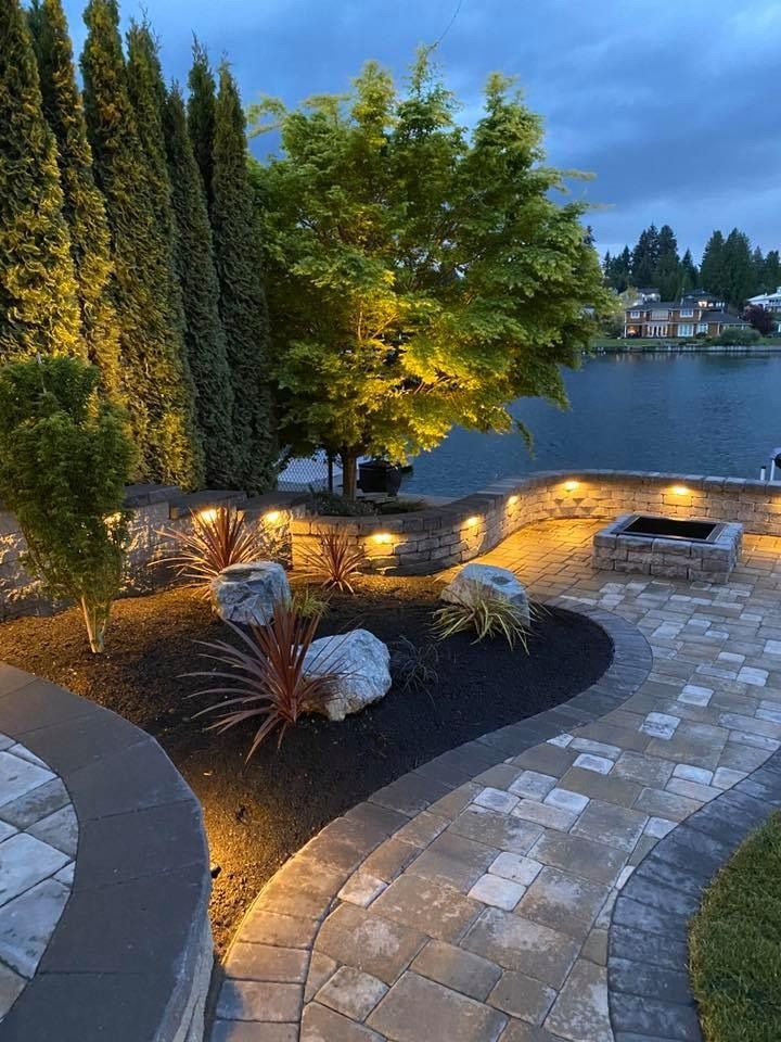Patio with up-lighting on trees and stone wall. Includes fire pit, lake, and patterned stone walkway.