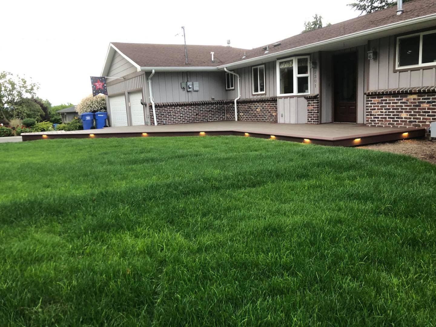 Lush green lawn slopes up to a concrete deck with recessed lights; a single-story house with a garage sits in the background.