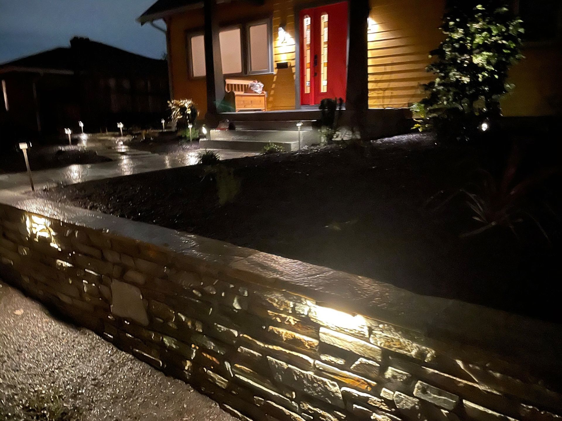 Night scene: a house with a lit red door, pathway and stone wall illuminated by outdoor lights.
