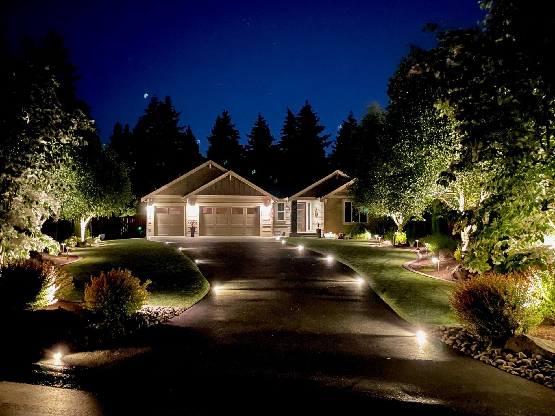 Nighttime view of a house with landscape lighting illuminating the driveway, lawn, and facade.