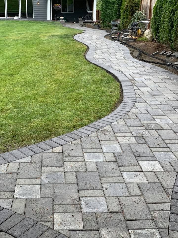 Curving paved walkway with grass border. Gray bricks, lush green lawn, and landscaping.