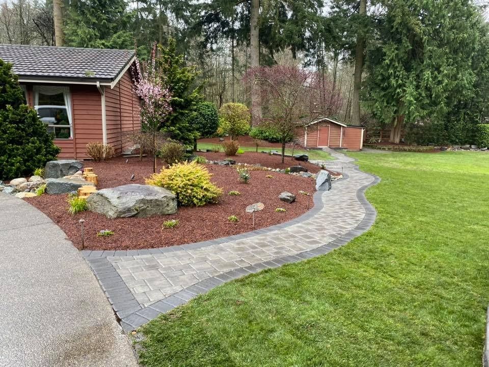 Brick pathway curves through a garden with trees, bushes, and a small building, surrounded by green grass.