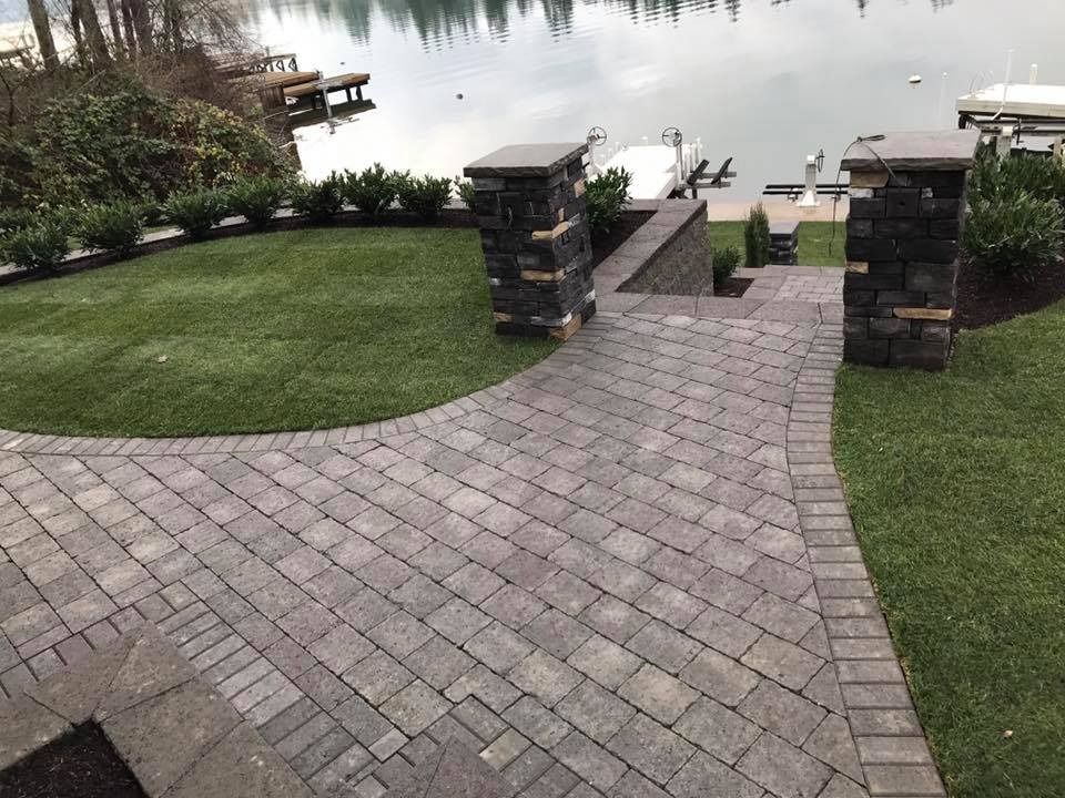 Brick pathway leading to a lake, flanked by green grass and stone columns.