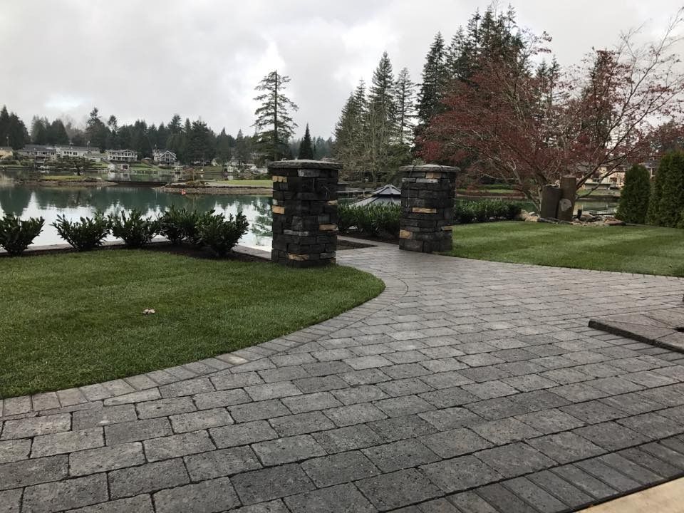 Brick pathway leading to a lake, flanked by green lawn, stone pillars, and trees under a cloudy sky.