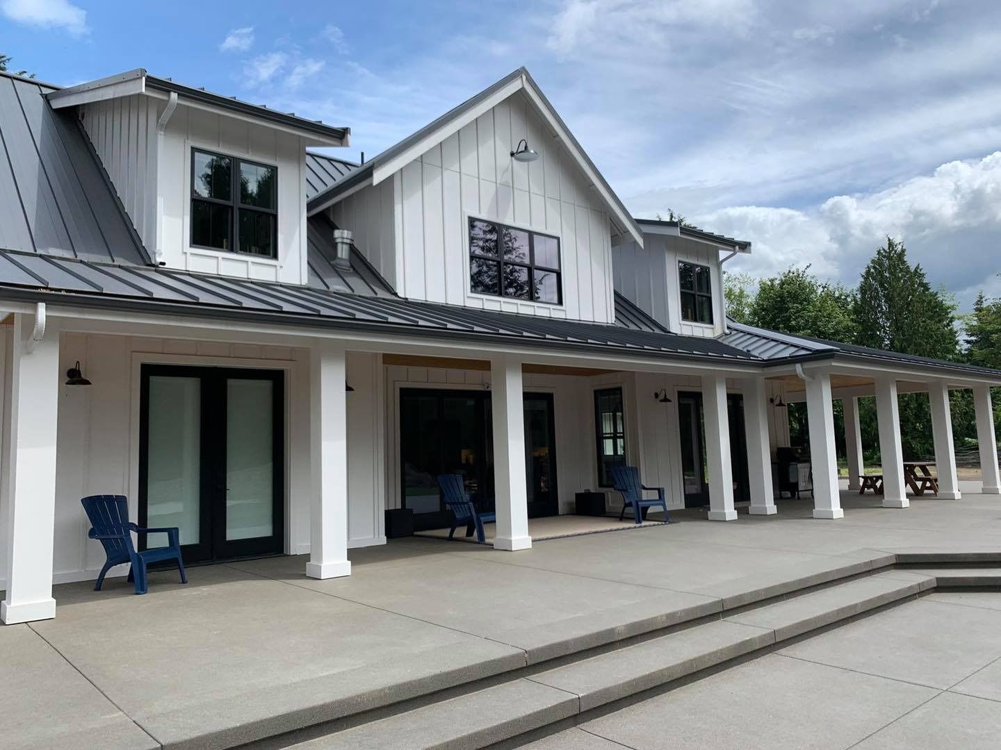 White farmhouse with dark roof, black windows, and a large porch, blue chairs on the patio.