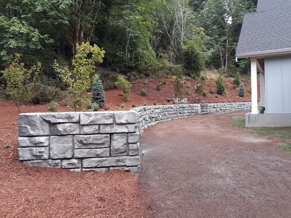 Stone retaining wall curves along a gravel driveway next to a building, with trees in the background.