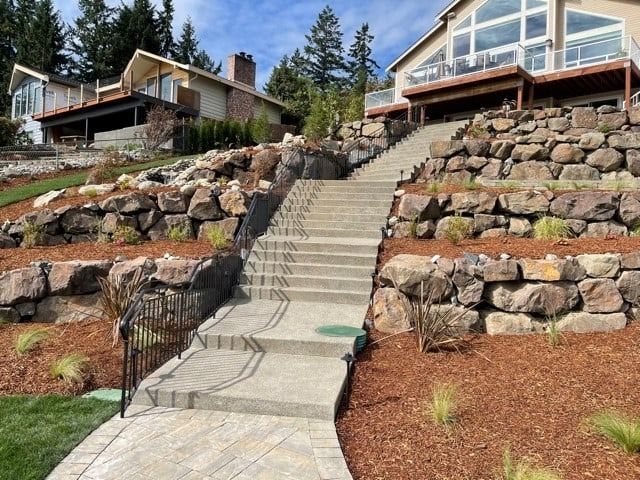 Stone staircase and retaining walls leading up to two houses on a hillside, with red mulch and green grass.