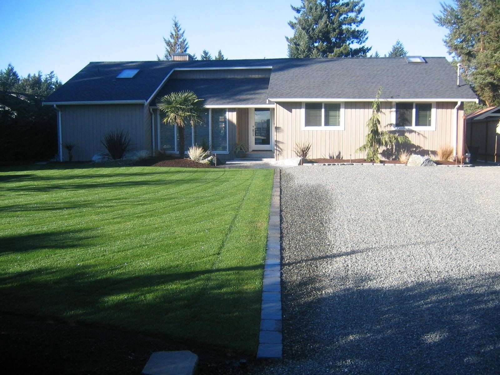 Ranch-style house with green lawn and gravel driveway; blue-grey trim, tan siding, blue sky.
