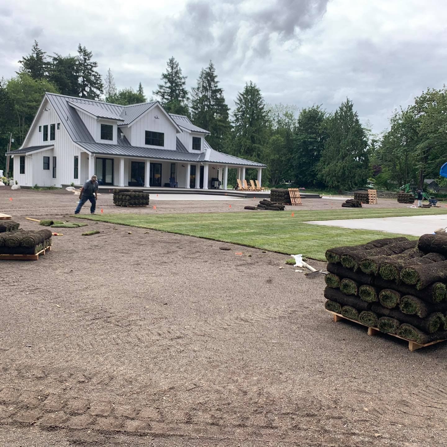 A person laying sod in front of a white farmhouse under a cloudy sky. Pallets of sod are visible.