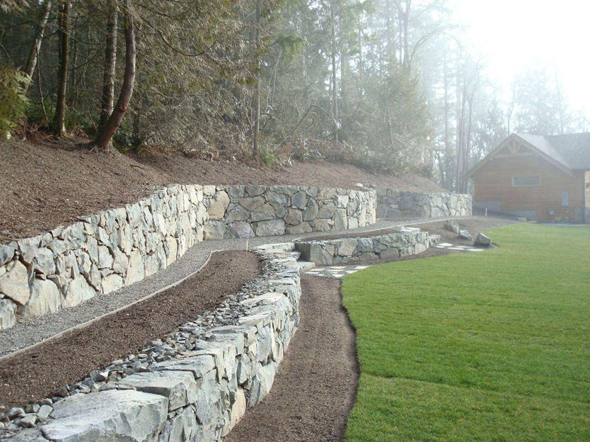 Stone retaining walls terraced into a hillside, bordering a lawn and forest.