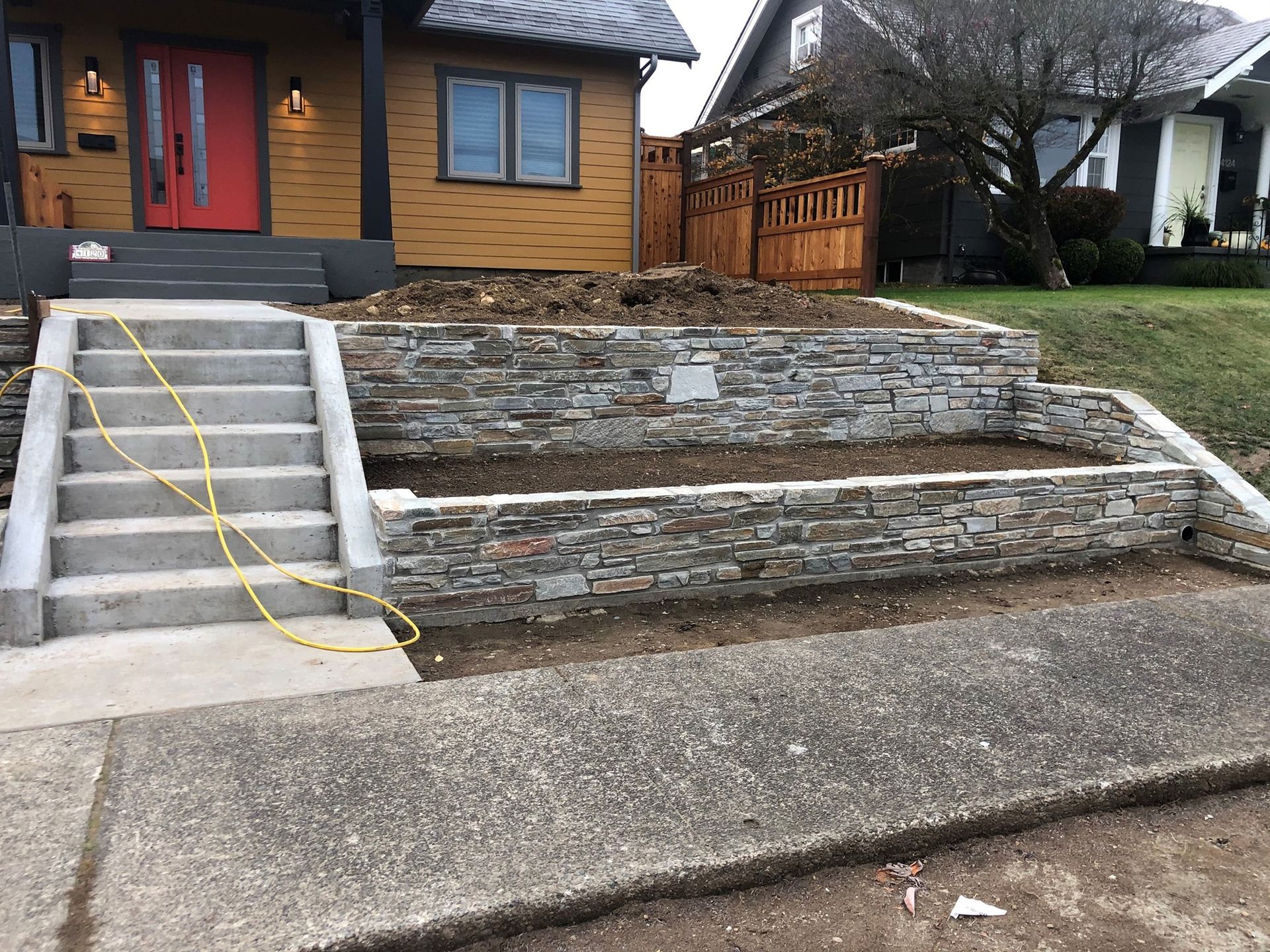 Exterior view of a house with gray stone retaining walls and concrete stairs; a yellow electrical cord is running along the stairs.