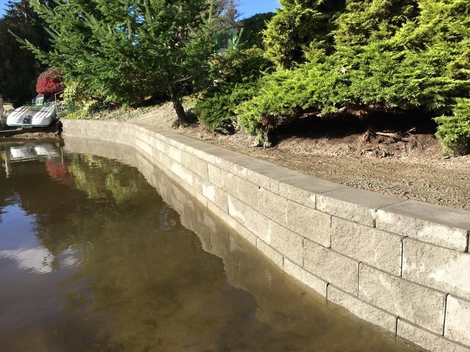 Stone retaining wall along the edge of a pond, with greenery and a blue sky reflected in the water.