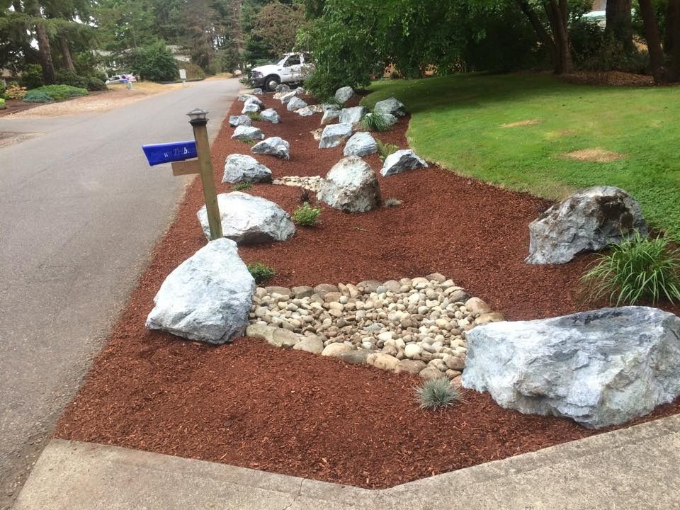 A roadside landscaped bed with mulch, rocks, small plants, and a rock-lined dry creek.