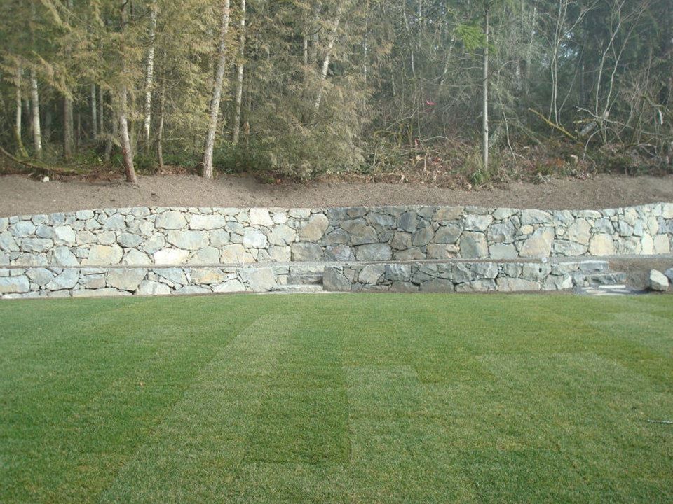 Stone retaining wall with green lawn and trees in the background.