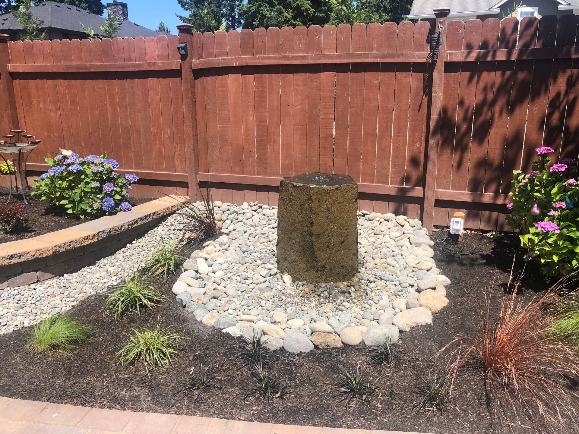 Water fountain in a rock bed in front of a stained wooden fence, surrounded by plants and flowers.