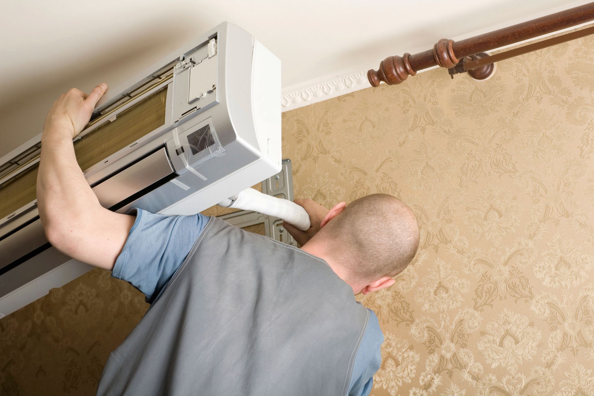 A professional technician installing a wall-mounted air conditioning unit in a room with patterned wallpaper.