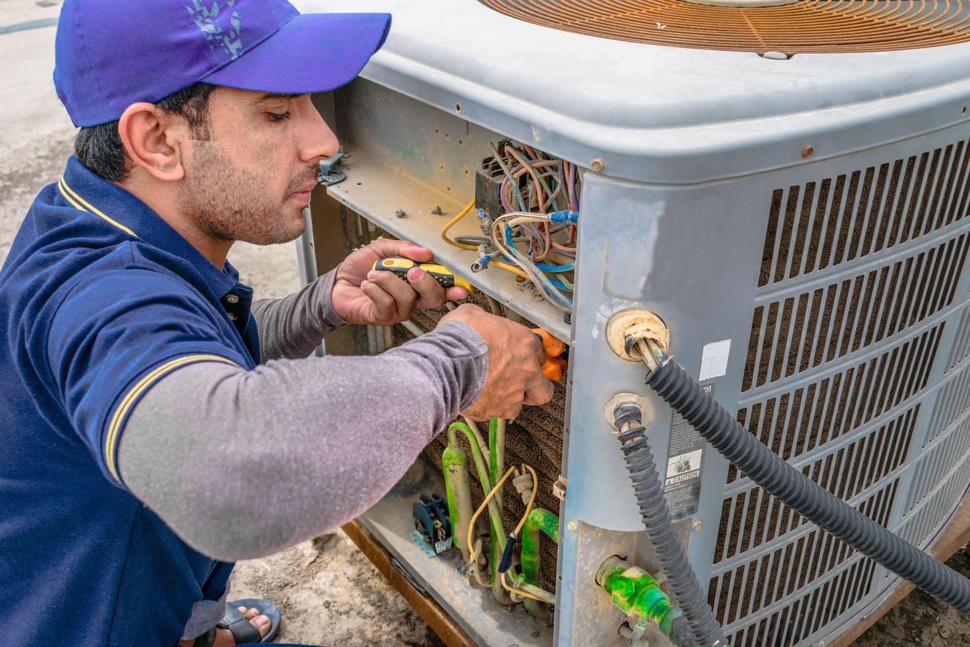 A technician in a blue shirt and hat repairs the wiring inside an outdoor air conditioning unit.