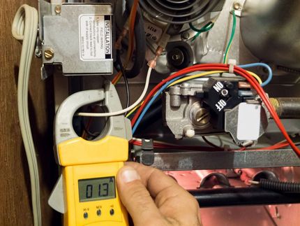 A hand uses a yellow clamp meter to measure the electrical current on a wire inside a gas furnace furnace control panel.