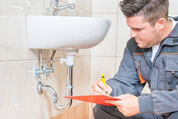 A plumber is looking at a sink in a bathroom and writing on a clipboard.