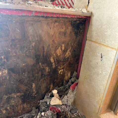 Damaged dark wall corner with peeling paint and debris on the floor beside a beige tiled wall.