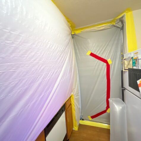 Plastic-covered hallway doorway sealed with gray sheeting and red tape, with yellow painter’s tape around edges