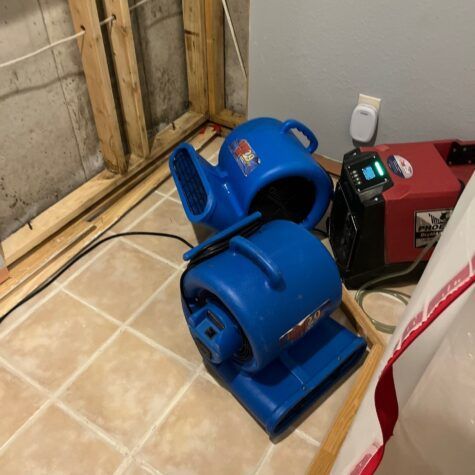Blue portable blower/fan on a tiled floor beside a red machine in a basement utility room
