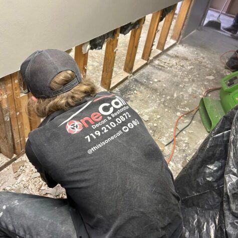 Worker removing drywall in a gutted room, viewed from behind, with exposed wall studs and construction debris.