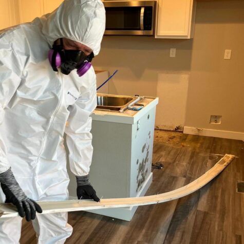 Worker in white hazmat suit and respirator inspecting damaged flooring in a room under renovation.