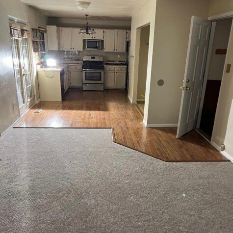 Small apartment kitchen with white cabinets and stainless stove, seen from carpeted living room with wood floor edge.