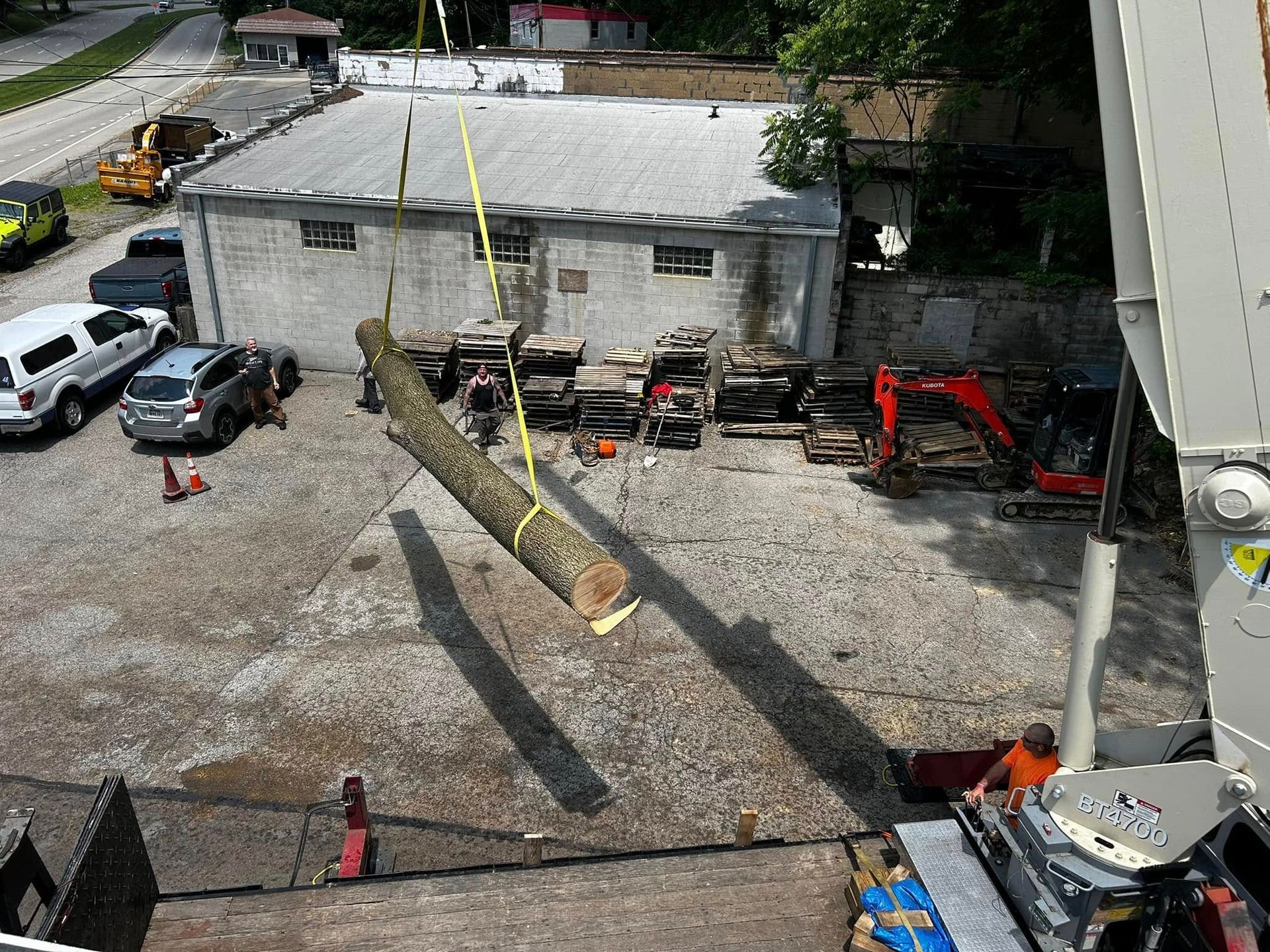 Construction site with a crane lifting a large tree trunk over a gravel lot near parked vehicles.