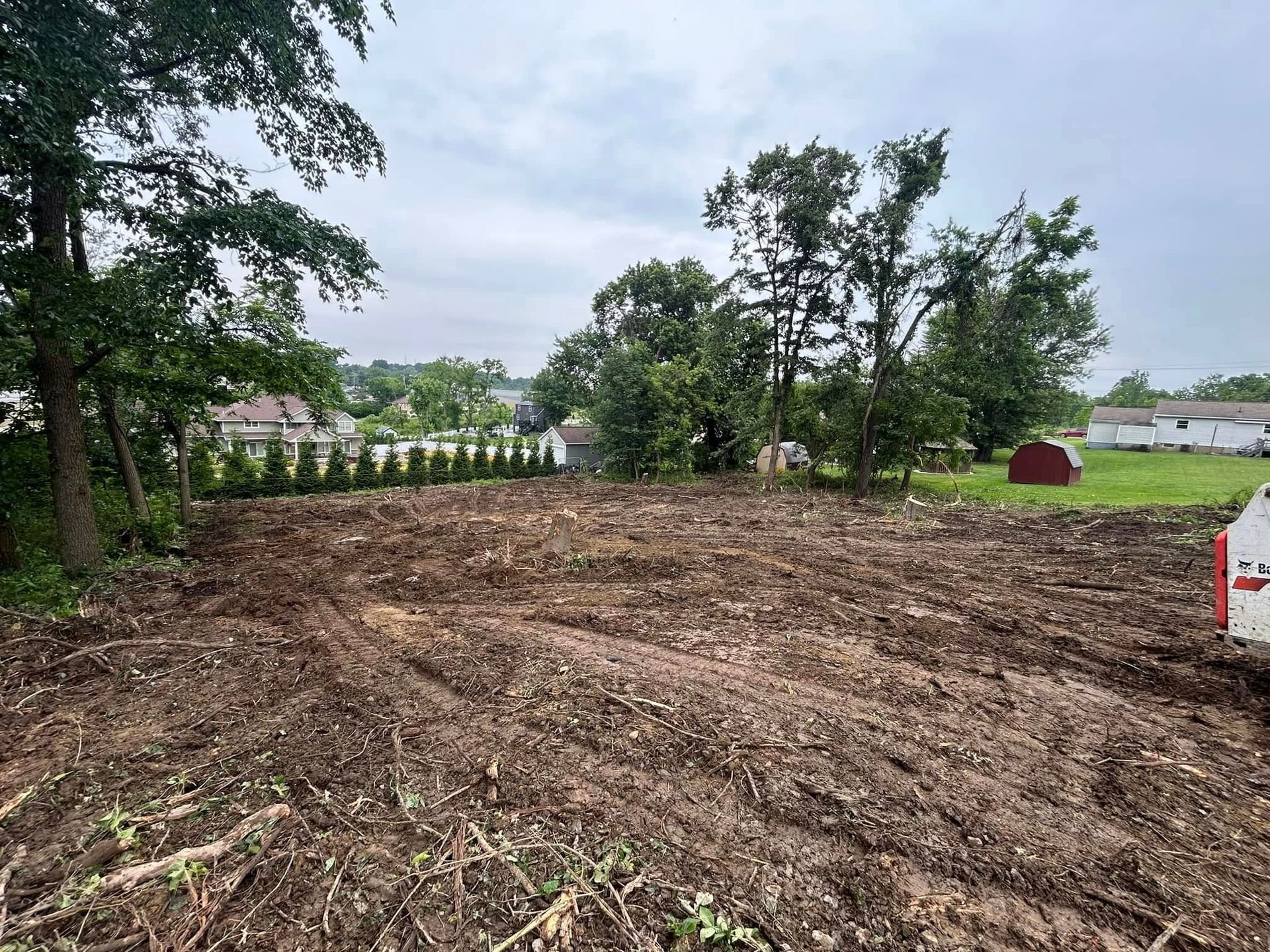 Cleared dirt lot with trees and a white fence under an overcast sky