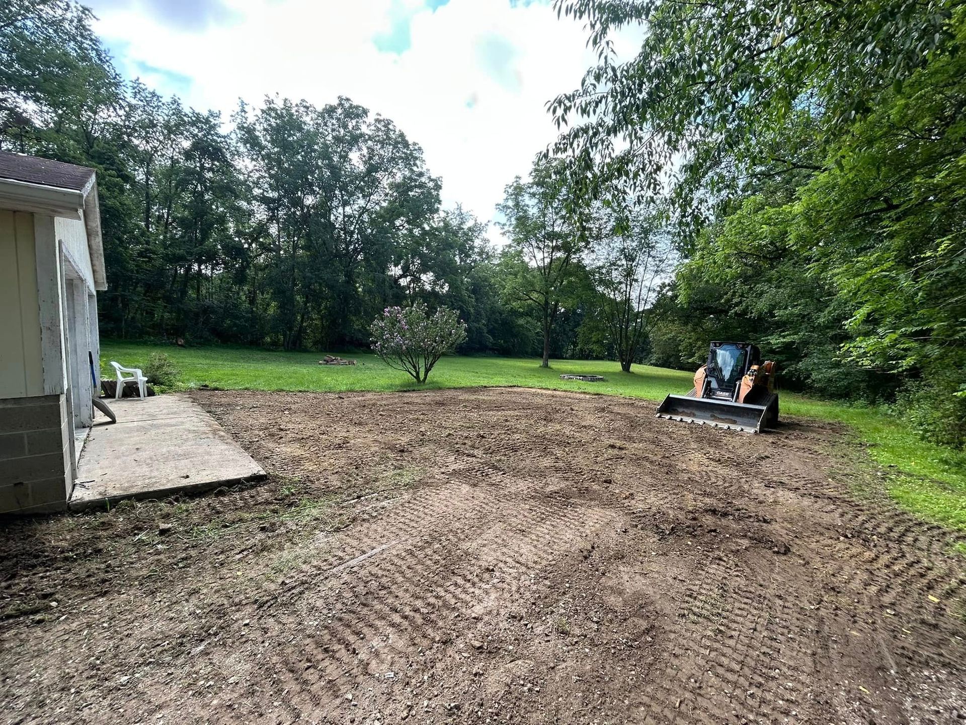 Cleared backyard with dirt ground, trees, and two people sitting on an ATV near the right side.