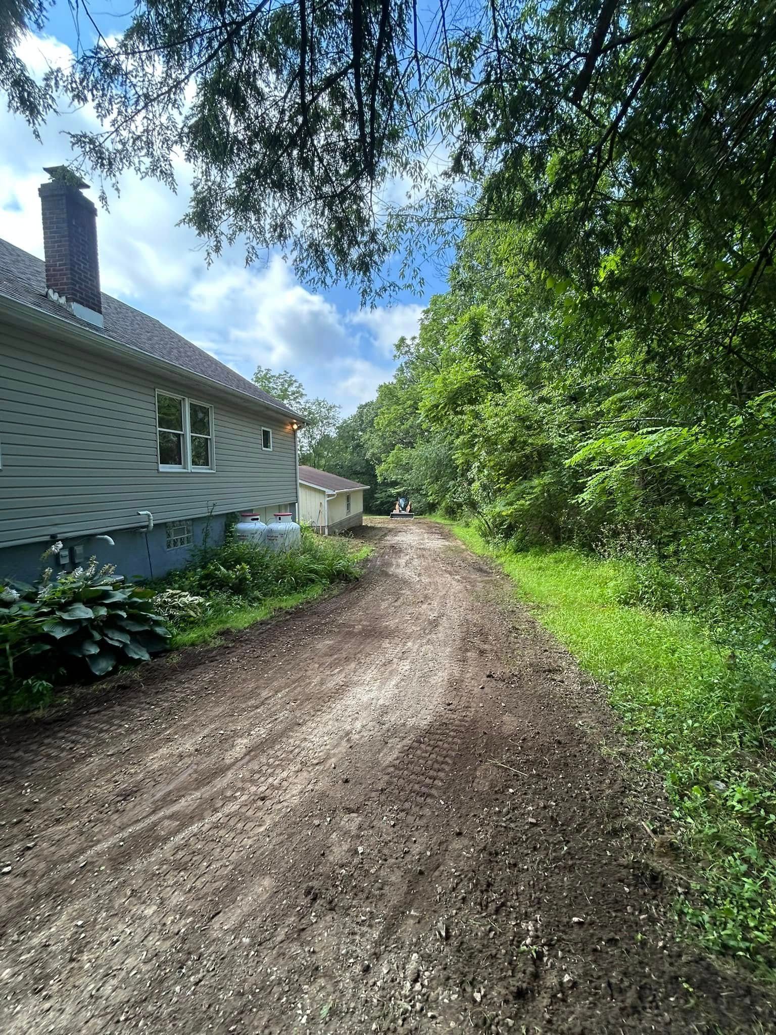 Dirt driveway beside a white house and leafy trees under a bright blue sky