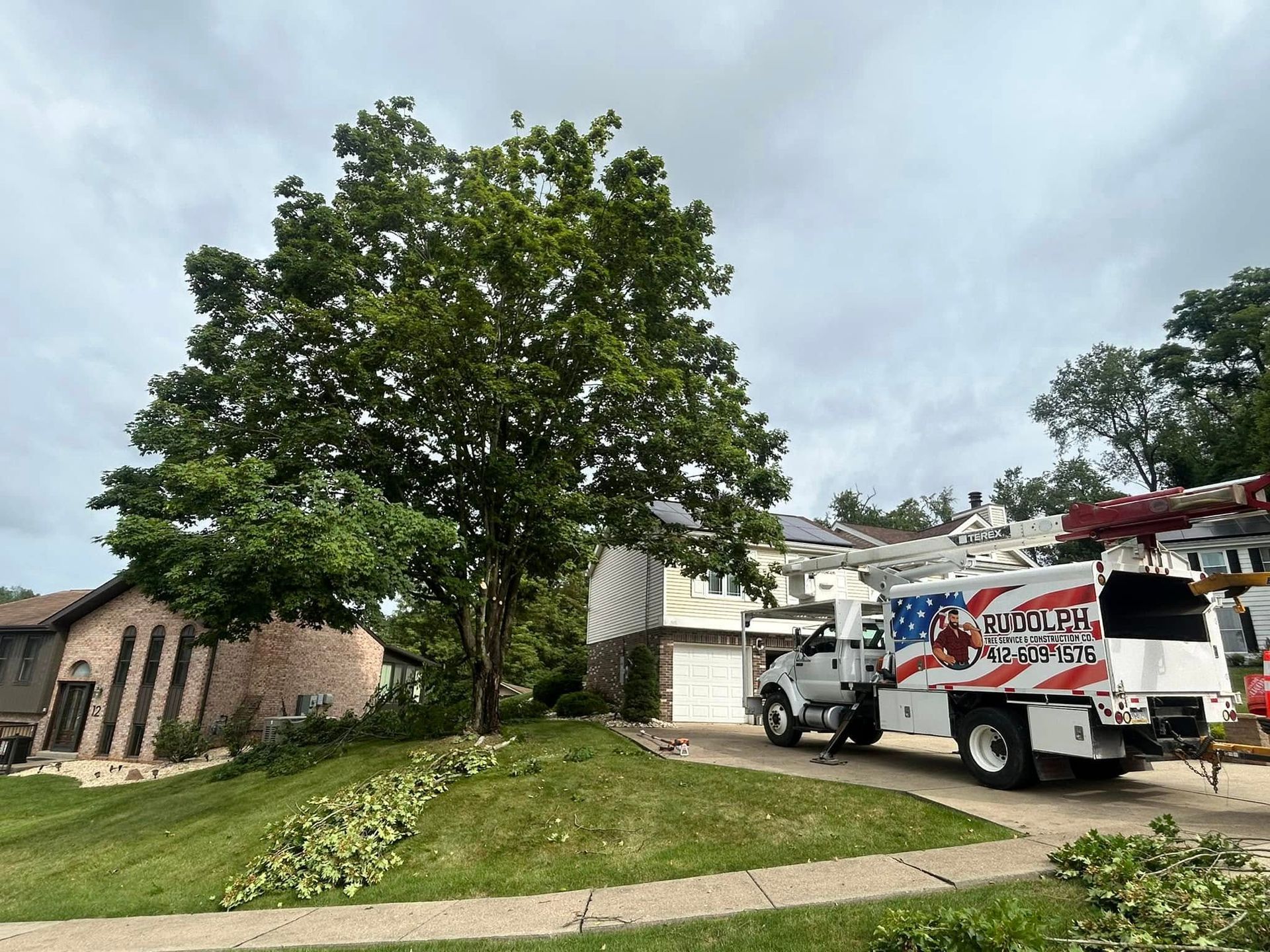 Utility truck and worker near a house, beside a large tree and grassy front yard under cloudy sky