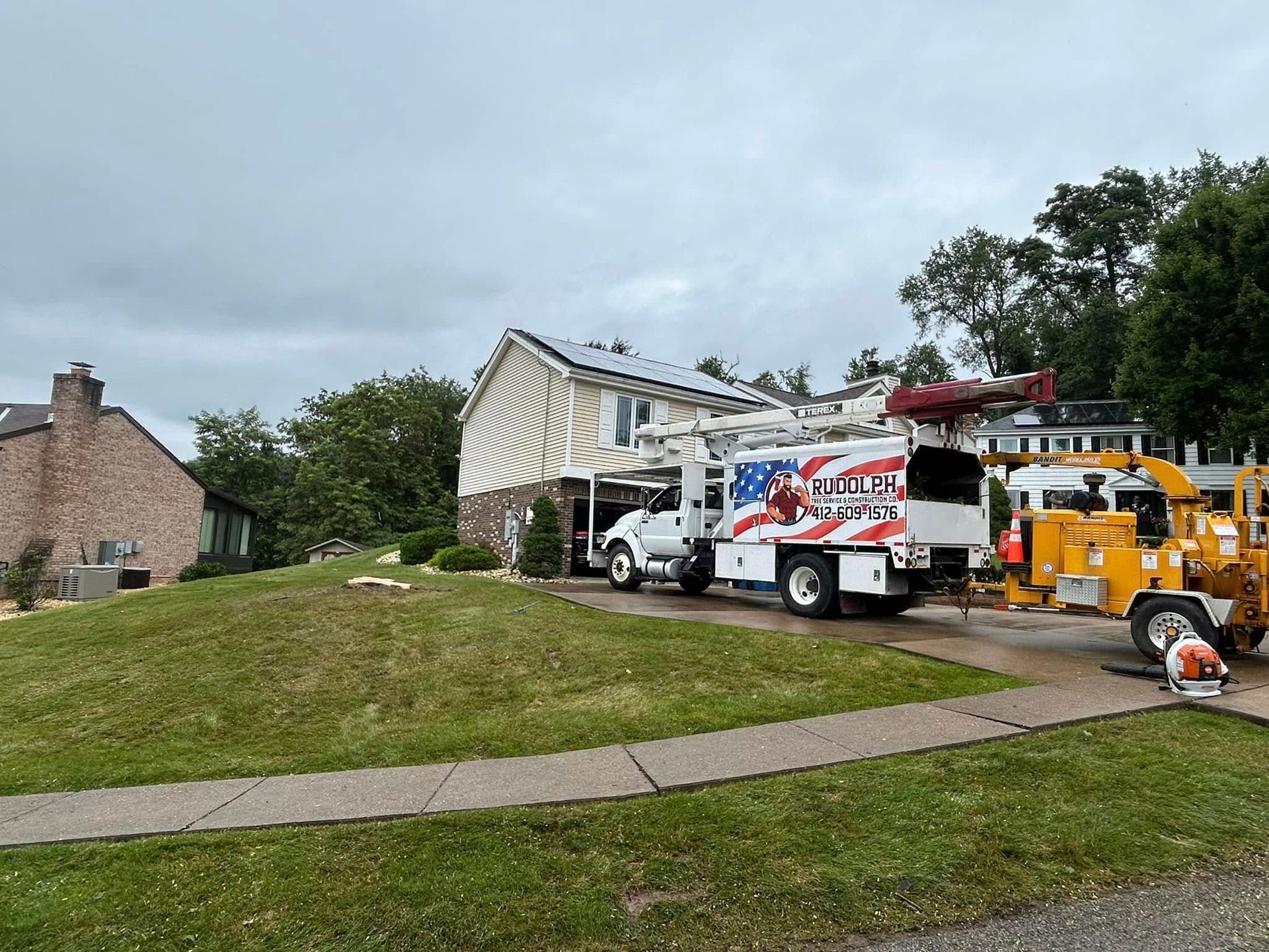Utility trucks and workers repairing storm damage beside a suburban house and fallen tree.