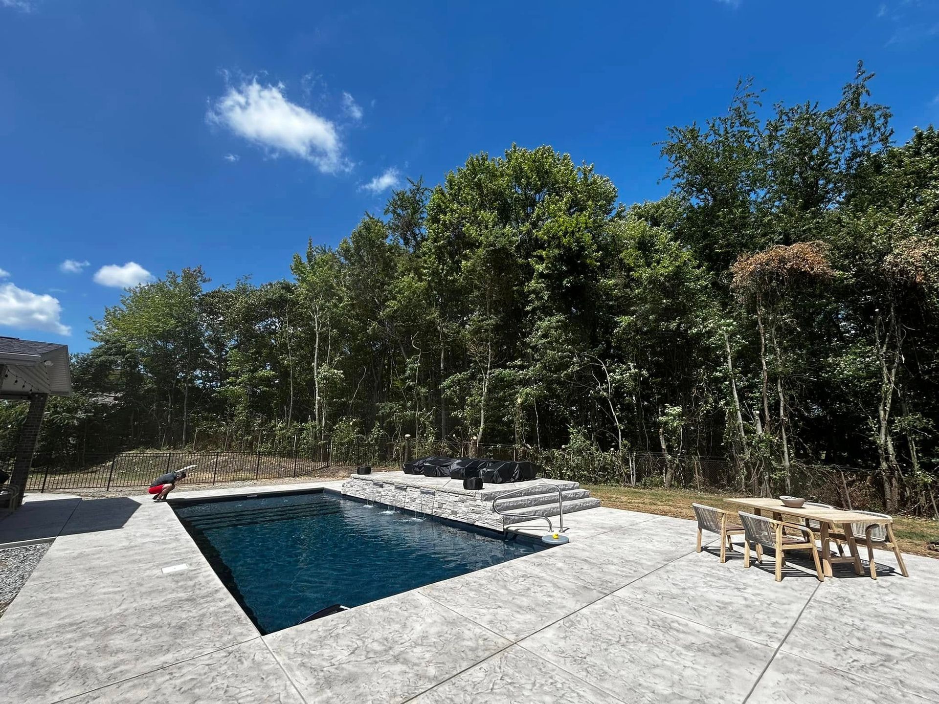 Backyard pool beside a concrete patio and wooded trees under a bright blue sky