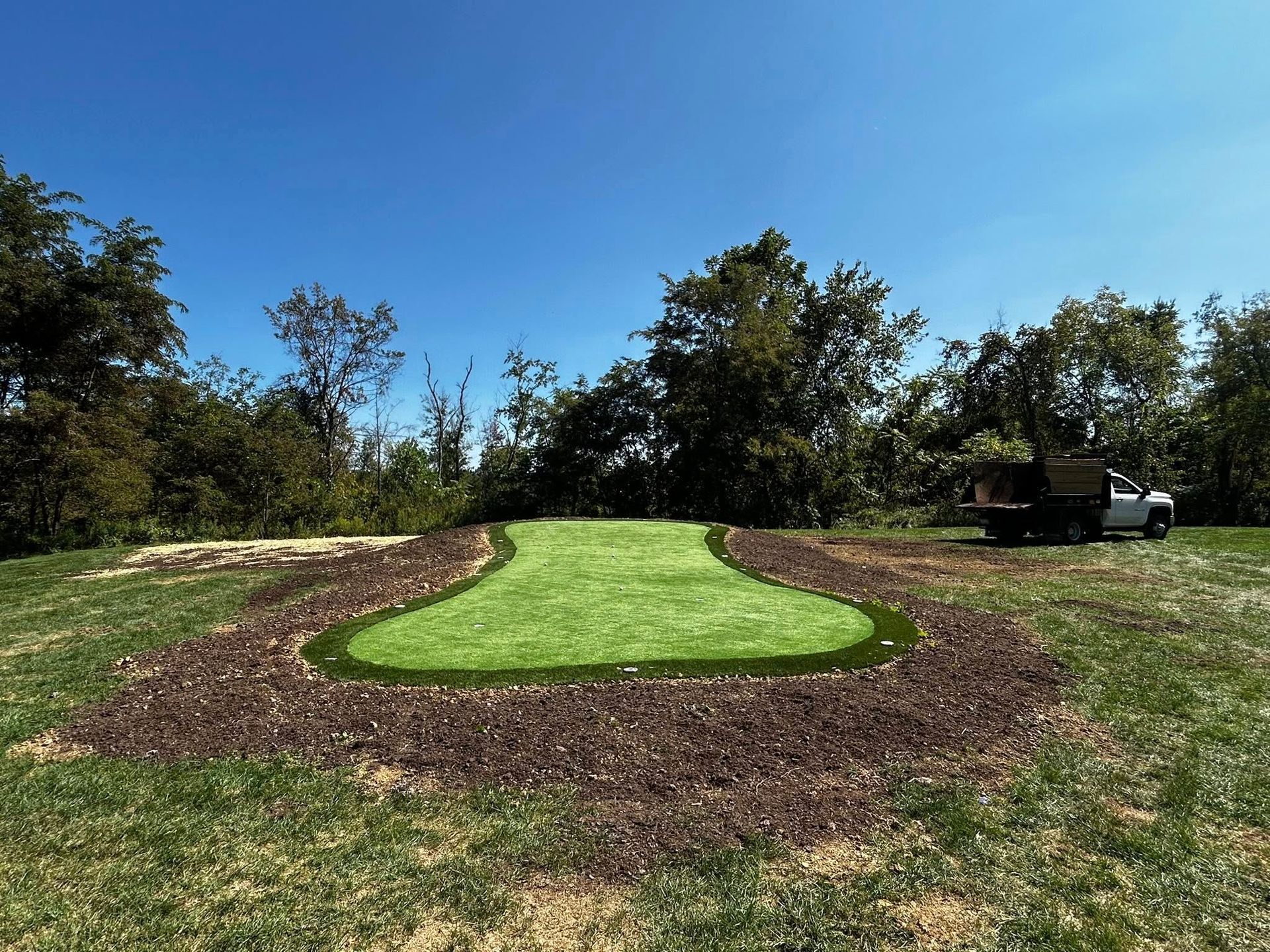 Mowed grass path bordered by mulch in a sunny park with trees and a parked truck in the background