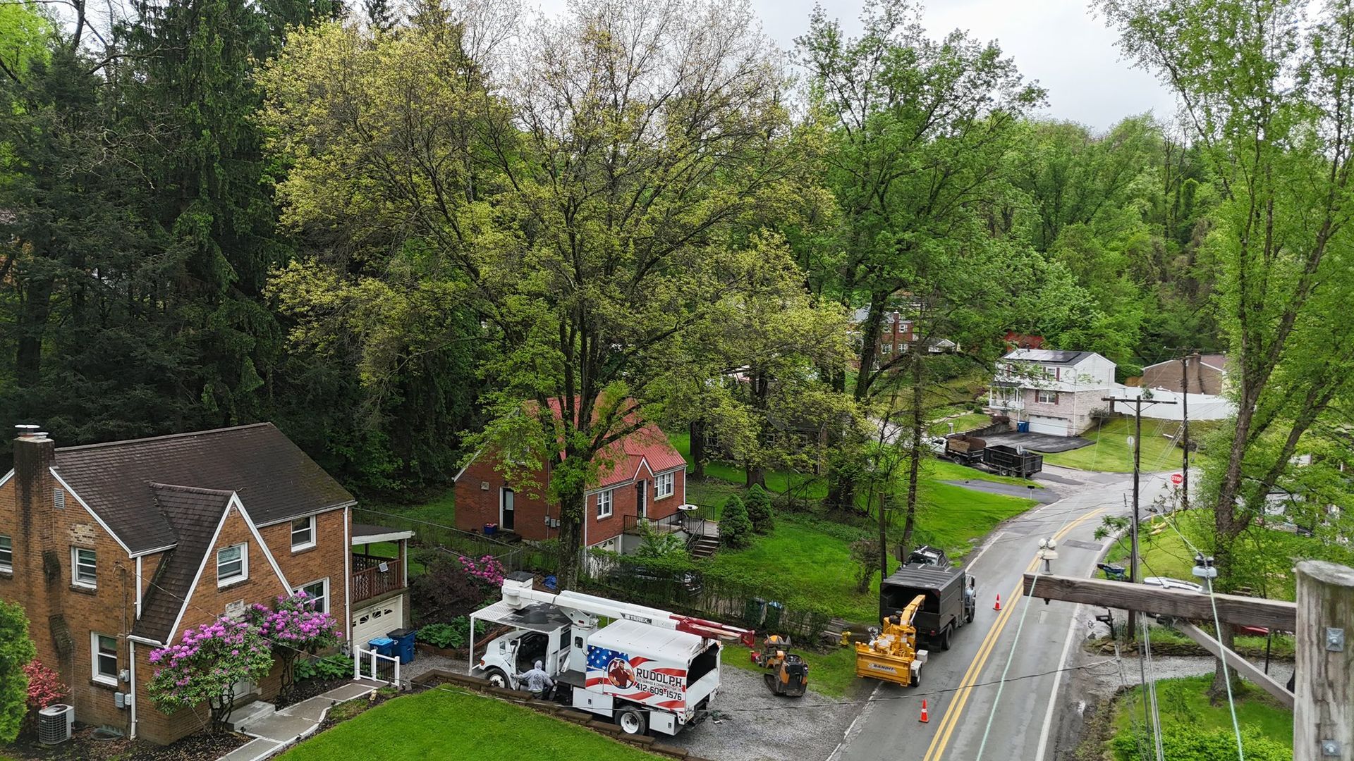 Aerial view of a rainy suburban street with homes, trees, parked trucks, and a road crew working by a roadside sinkhole