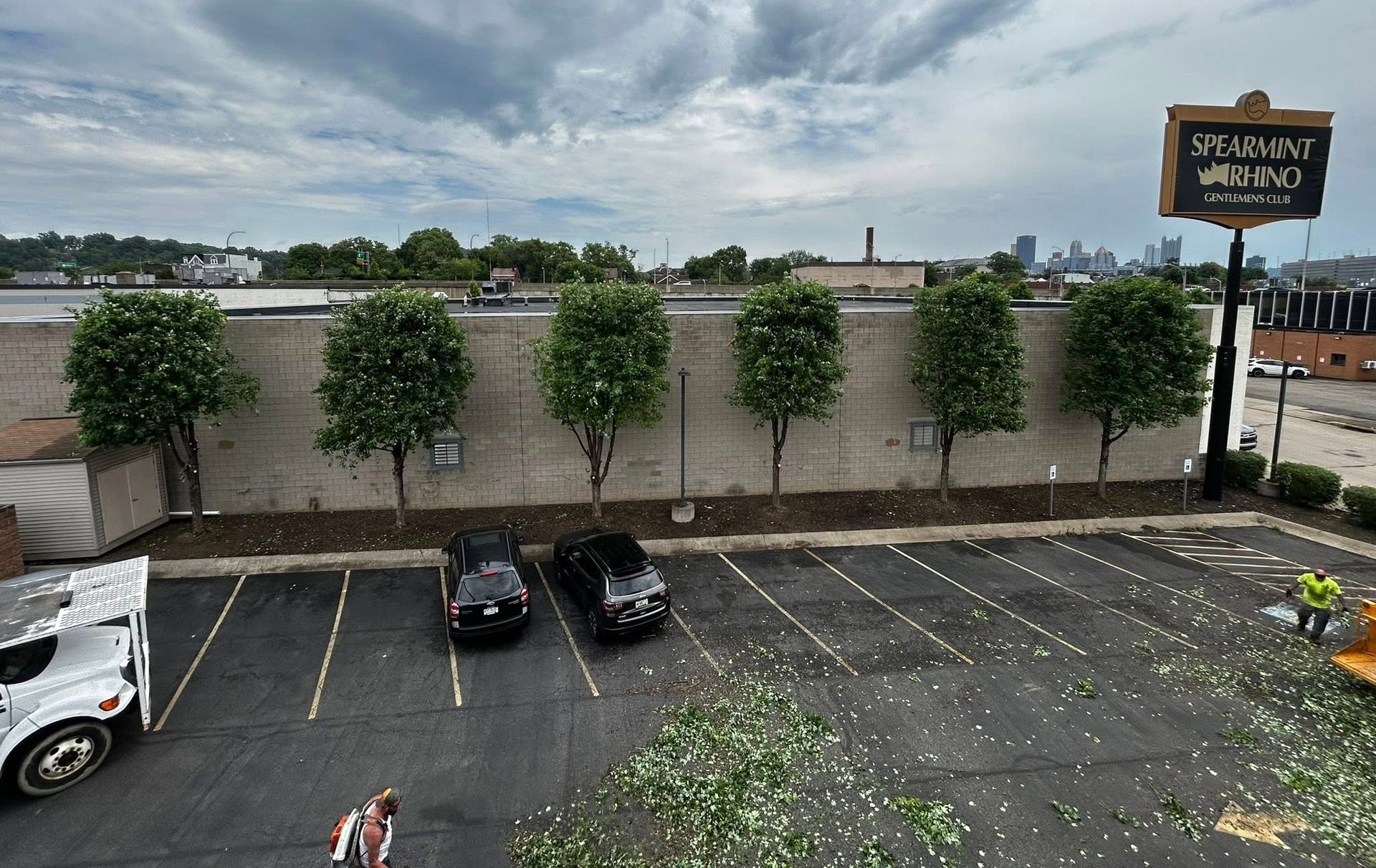 Parking lot beside a stone wall with trees under a cloudy sky, with cars parked below a sign.